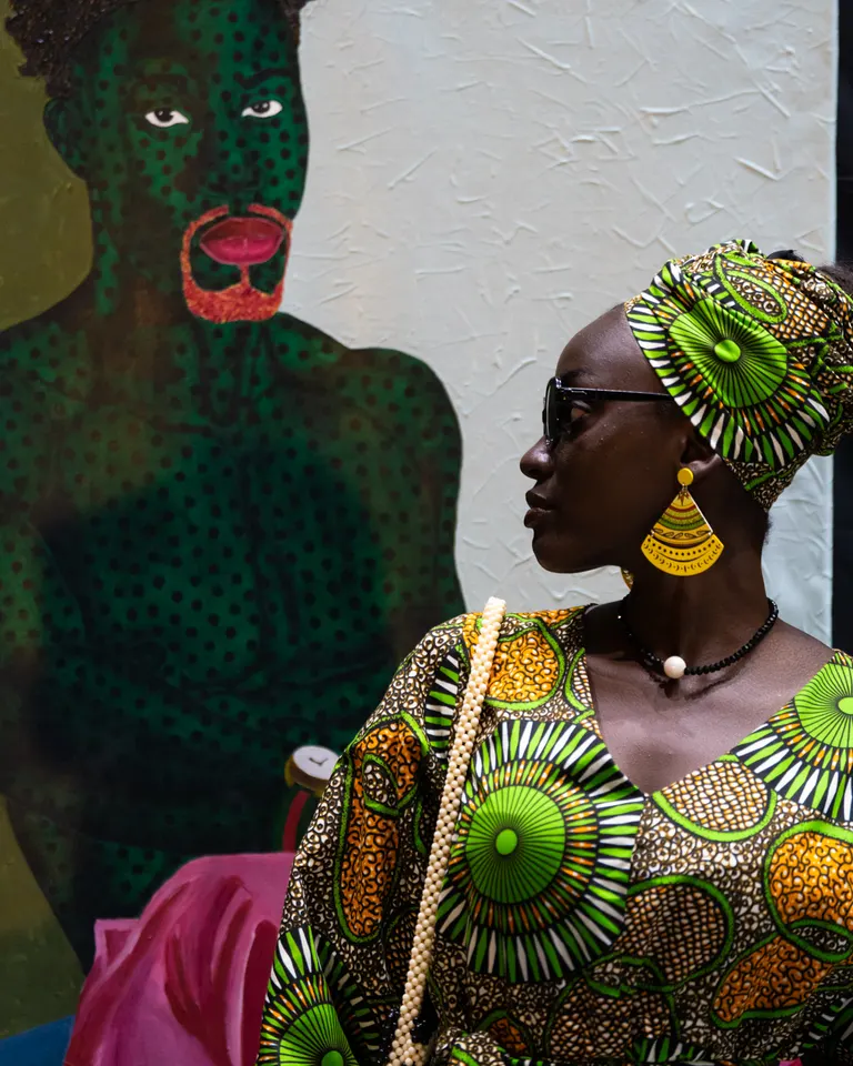 A visitor in a patterned outfit beside a painted portrait at KAFART 2024.